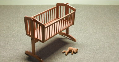 A baby cradle sits empty with a teddy bear on the floor next to it.