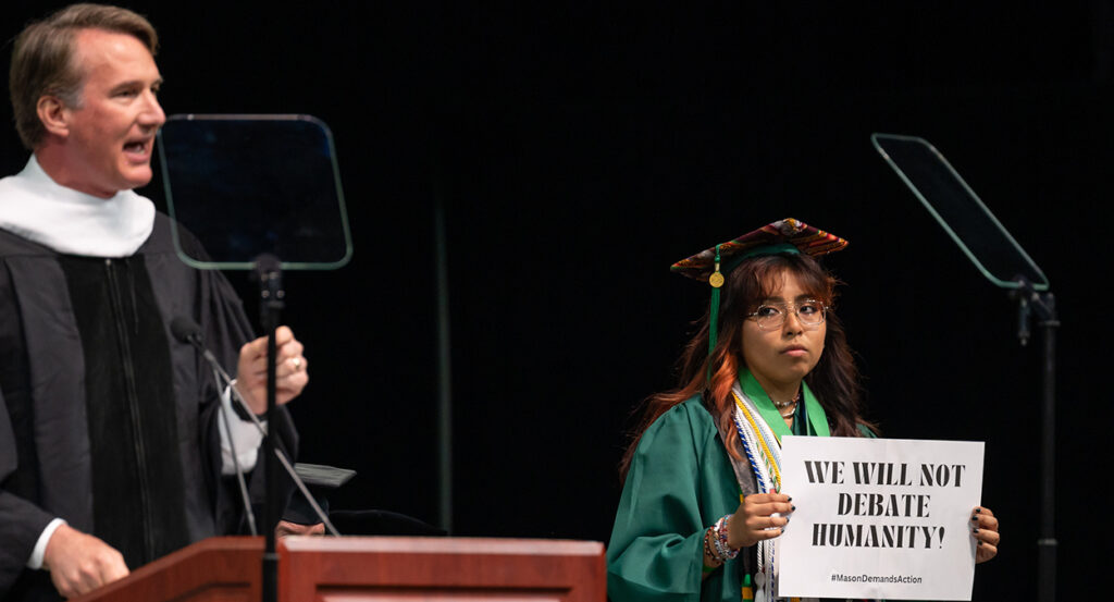 George Mason University graduate holds a sign reading "We will not debate humanity" while Gov. Glenn Youngkin speaks.