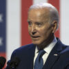 Joe Biden raises his finger in a blue suit with an American flag pin.