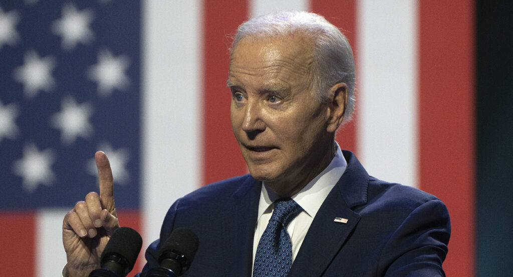 Joe Biden raises his finger in a blue suit with an American flag pin.
