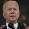 President Joe Biden gestures in front of a microphone while wearing a suit and a blue striped tie
