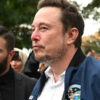 Elon Musk walks outside after leaving a meeting wearing a white shirt and dark blue jacket.