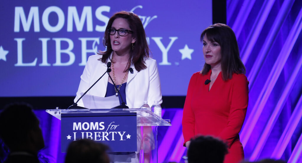 Tiffany Justice in a blue dress and white jacket stands next to Tina Descovich in a red dress in front of a Moms for Liberty sign.