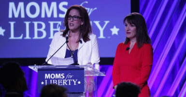 Tiffany Justice in a blue dress and white jacket stands next to Tina Descovich in a red dress in front of a Moms for Liberty sign.
