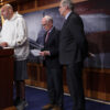 John Fetterman stands behind a congressional podium wearing a white hoodie, grey shorts and grey running shoes. Bernie Sanders, Ed Markey, Peter Welch, and Jeff. Merkley stand behind him.