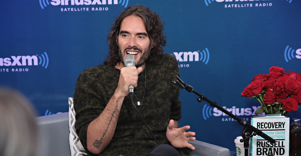 Russell Brand sits wearing a black shirt and speaks into a microphone.