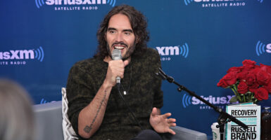 Russell Brand sits wearing a black shirt and speaks into a microphone.