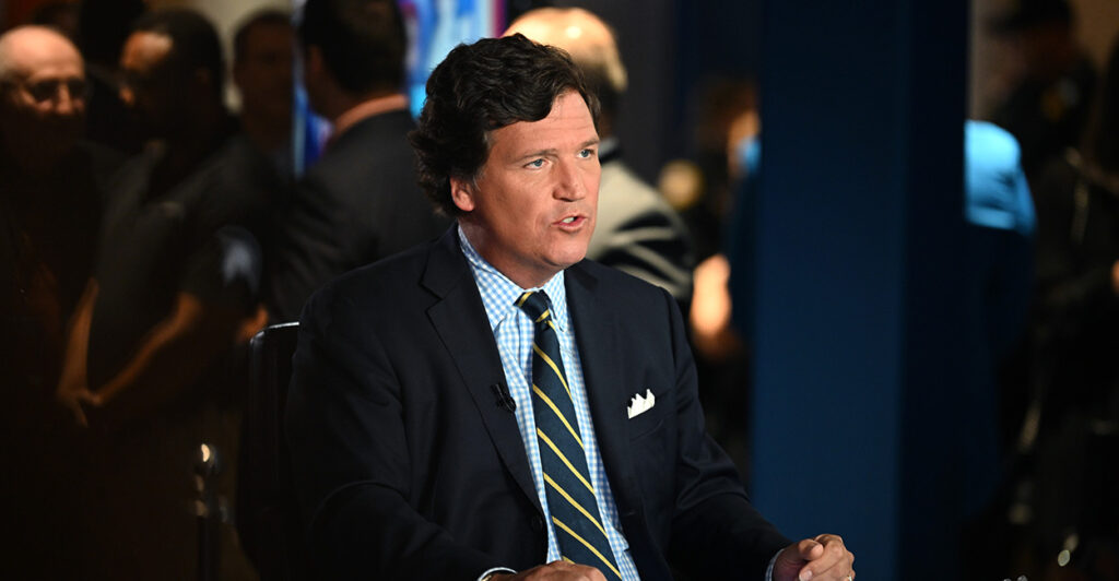 Tucker Carlson sits wearing a black suit and a blue shirt with a tie.