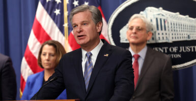 FBI Director Christopher Wray stands at a podium with Attorney General Merrick Garland and Assistant Attorney General Lisa Monaco behind him