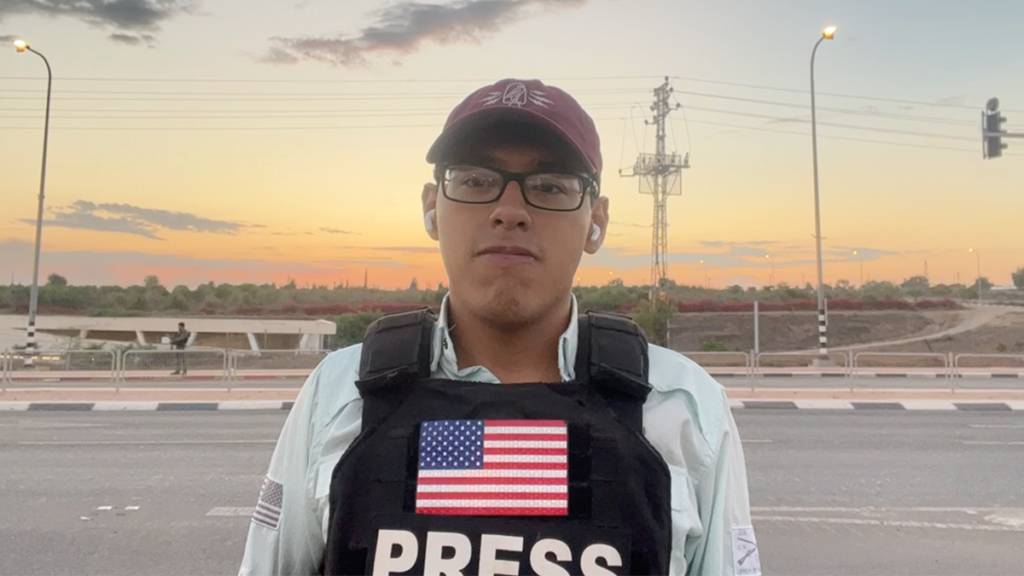 Reporter Julio Rosas outside with a baseball cap and a bulletproof vest with a U.S. flag and the word press across the chest