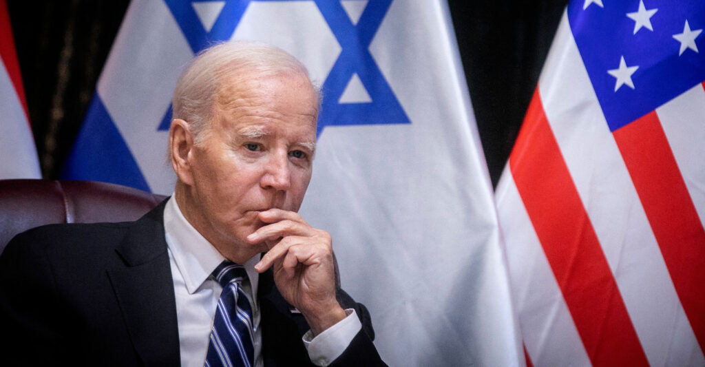 President Biden in a suit leans in front of an Israeli flag and an American flag