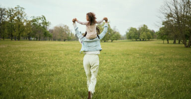 Mother walks through a field with her young daughter on her shoulders.