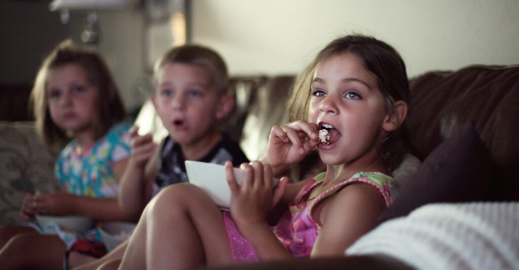 From left to right, a girl, a boy, and a girl sit on a couch eating popcorn