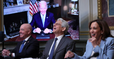 Alejandro Mayorkas, Merrick Garland, and Kamala Harris sit together laughing. Joe Biden smiles on a screen behind them.