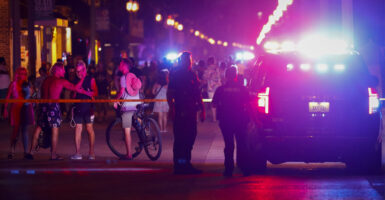 Beachgoers are seen next to law enforcement officers on a crime scene at night with police cars with lights flashing.