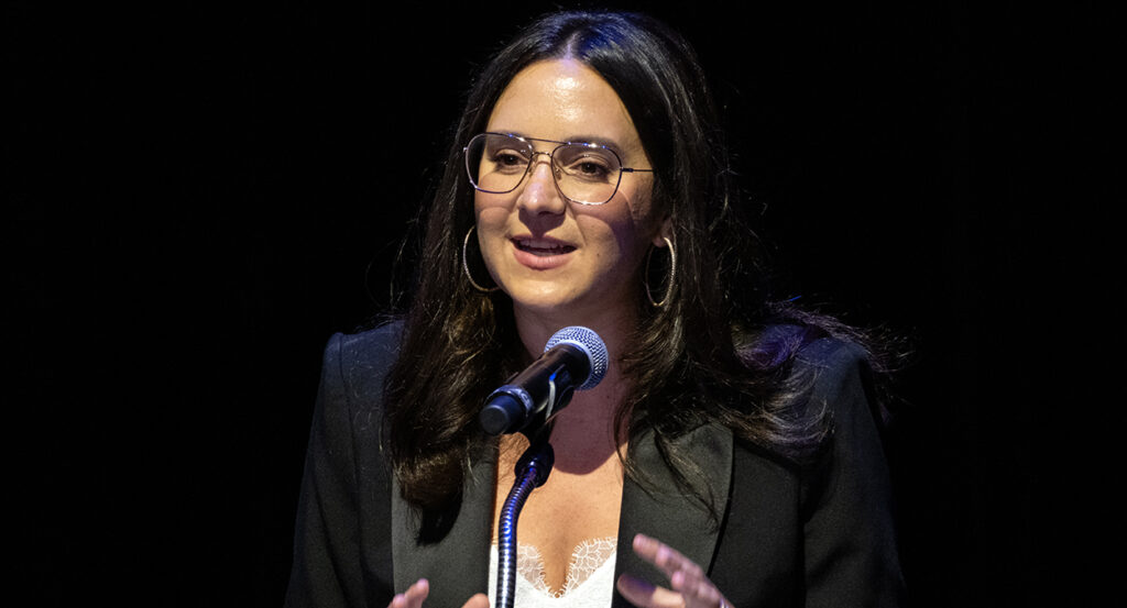 Bari Weiss in a white dress and a black blazer speaks in front of a microphone