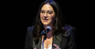 Bari Weiss in a white dress and a black blazer speaks in front of a microphone