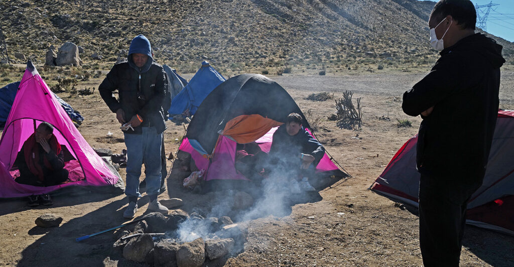A few small tents are seen around a campfire where two illegal aliens stand.