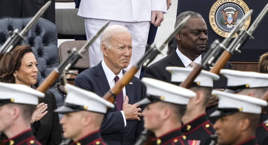Kamala Harris, Joe Biden, and Lloyd Austin in suits stand in front of soldiers carrying rifles with bayonets.