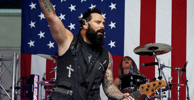 Skillet's John Cooper raises his hand while playing guitar and singing in front of an American flag.
