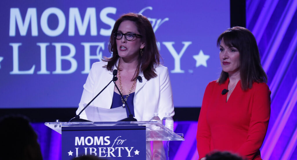 Tiffany Justice in a blue dress with a white suit jacket stands in front of a Moms for Liberty logo with Tina Descovich in a red dress right next to her