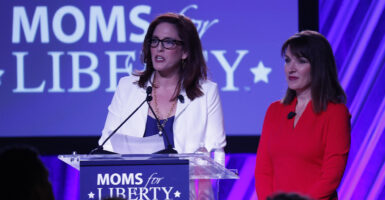 Tiffany Justice in a blue dress with a white suit jacket stands in front of a Moms for Liberty logo with Tina Descovich in a red dress right next to her
