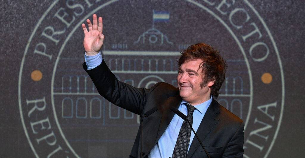 javier milei waves on stage in a black jacket and a blue shirt