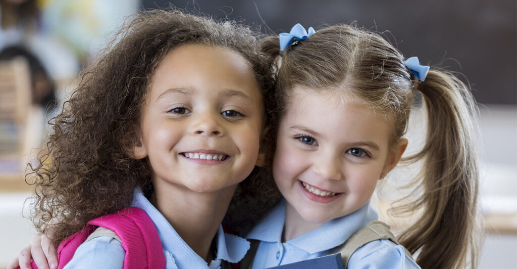 Two kindergarten girls wearing school uniforms hug each other in their classroom.