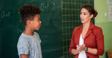 A white female teacher and black middle school student stand in front of a chalkboard with math equations