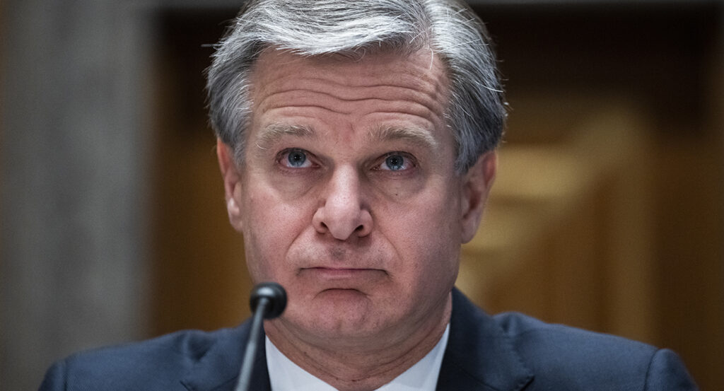 Christopher Wray in a blue suit speaks in front of a microphone