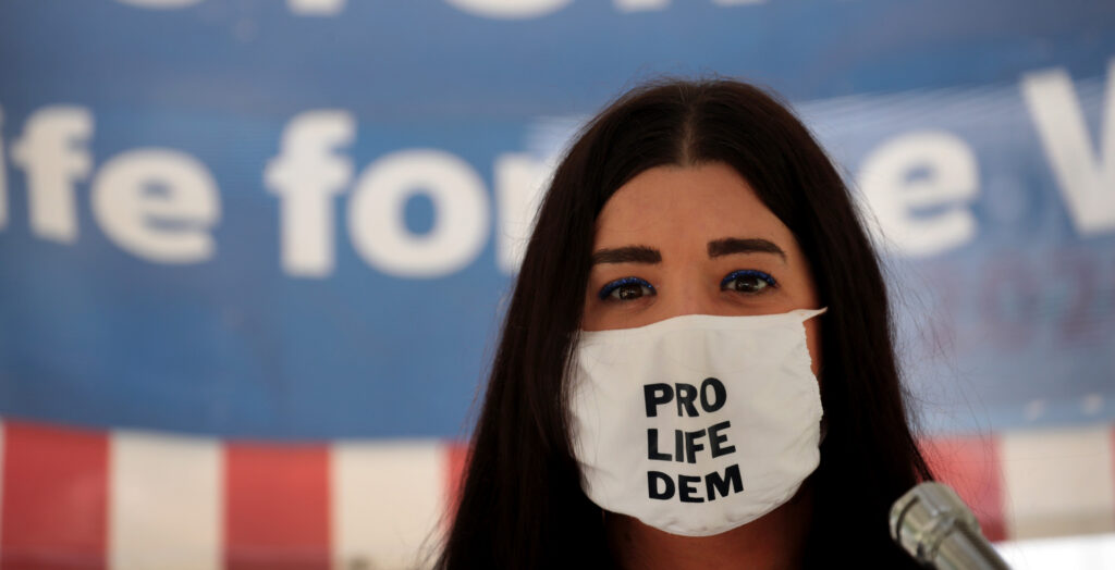 Terrisa Bukovinac stands in front of an American flag while wearing a face mask with the message "pro life Dem"