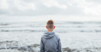 A teen boy stands staring at the ocean with his back to the camera.