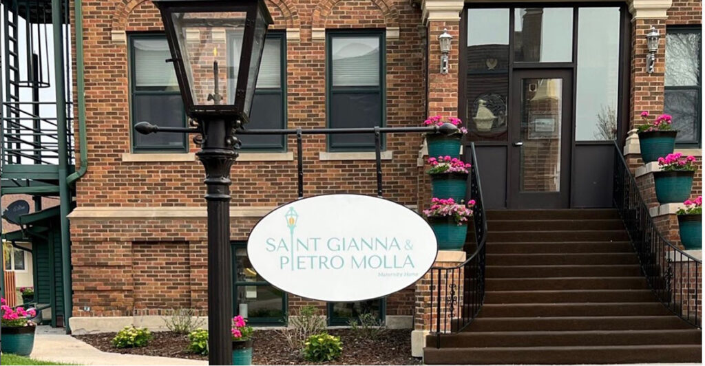 The St. Gianna and Pietro Molla Home sign hands on a lamp post outside the large brick home.