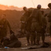 A group of Israeli troops organizes equipment on the ground as the sun peeks over a hill.