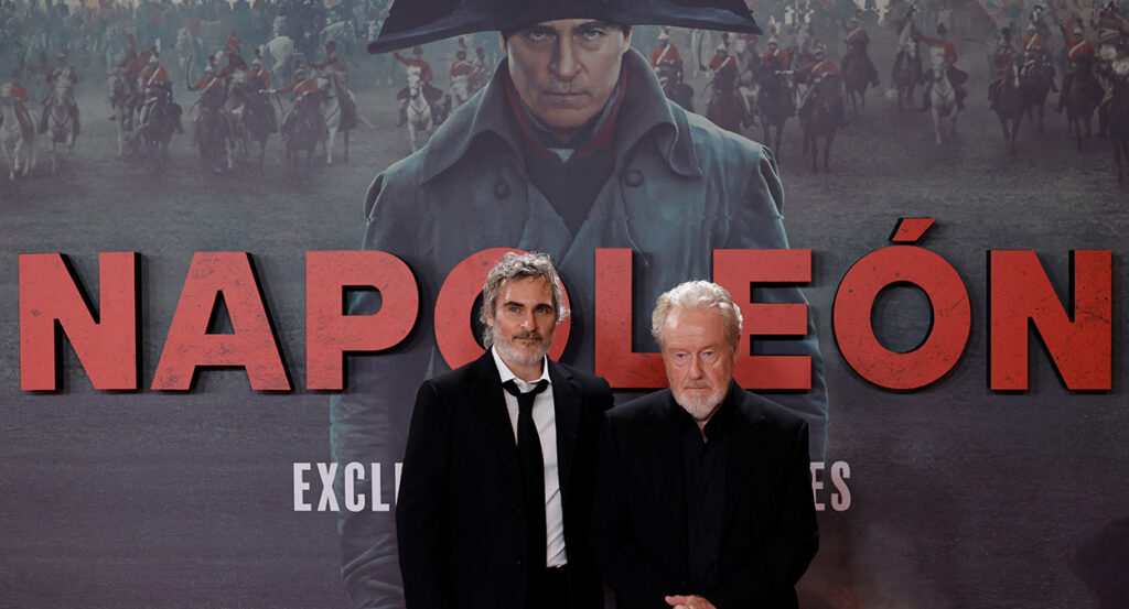 Joaquin Phoenix and Ridley Scott stand in front of a poster for the Napoleon 2023 movie