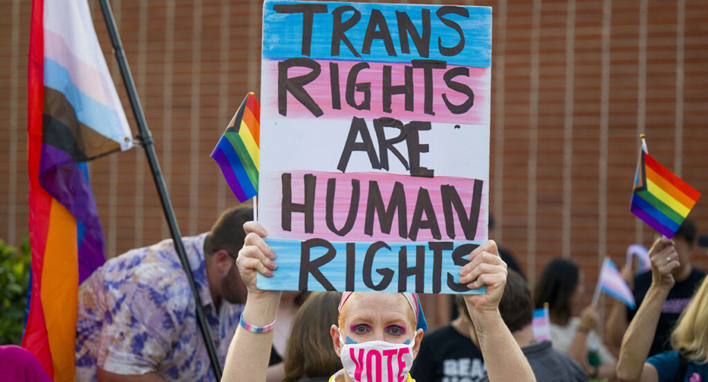 Woman holds sign reading "trans rights are human rights" while wearing a face mask reading "Vote"