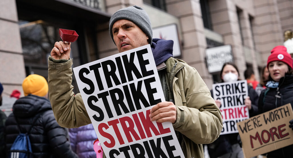 Man in jacket holds sign reading "Strike" while shaking a cowbell.