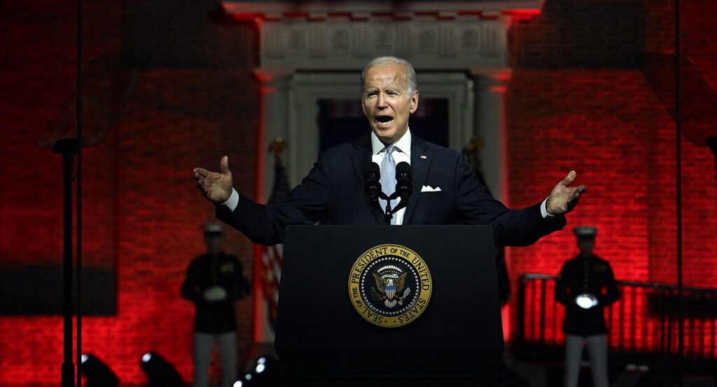 President Joe Biden gestures in a black suit in front of Philadelphia's Independence Hall behind the presidential seal