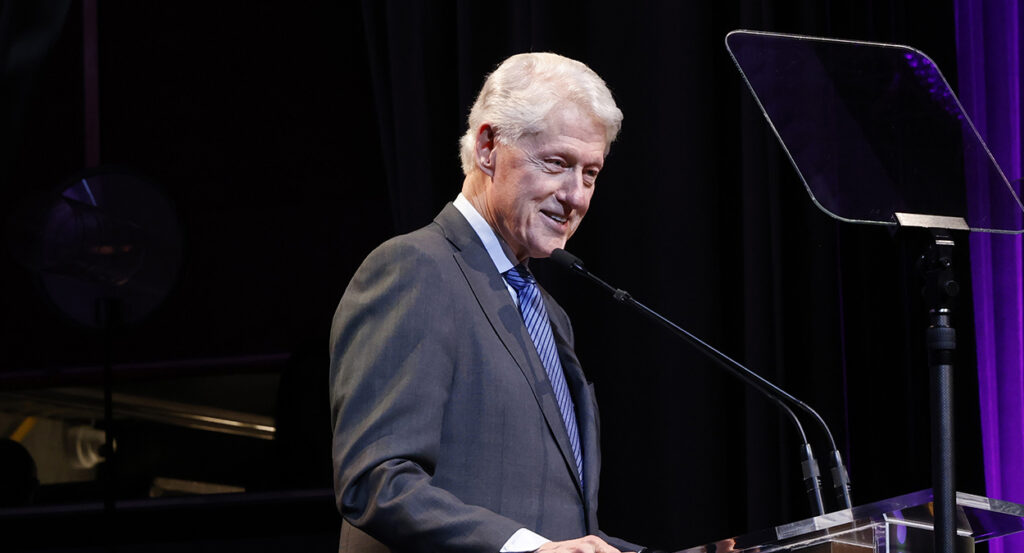 Bill Clinton in a grey suit speaks behind a teleprompter