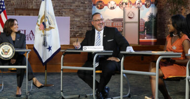 Kamala Harris in a suit, Miguel Cardona in a suit, sit behind desks