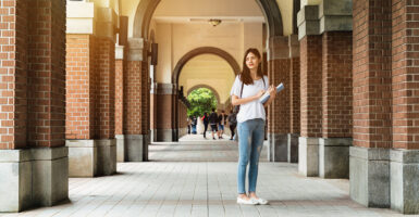 A girl stands in a passageway on a college campus with people in the distance behind her.