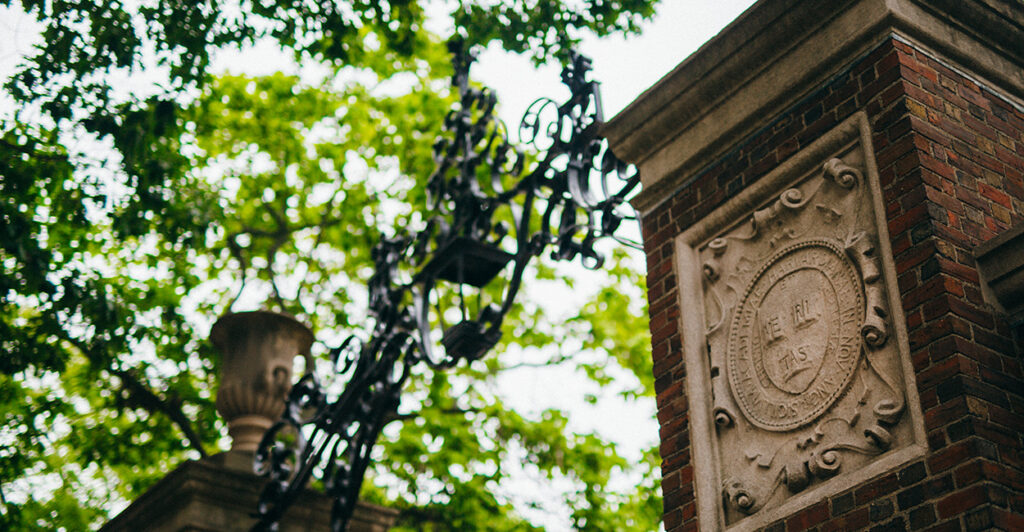 A brick and iron entry to Harvard is seen below a green tree on a sunny day.