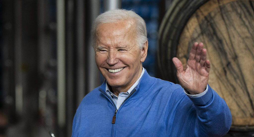 President Joe Biden waves in a blue sweatshirt in front of a beer barrel