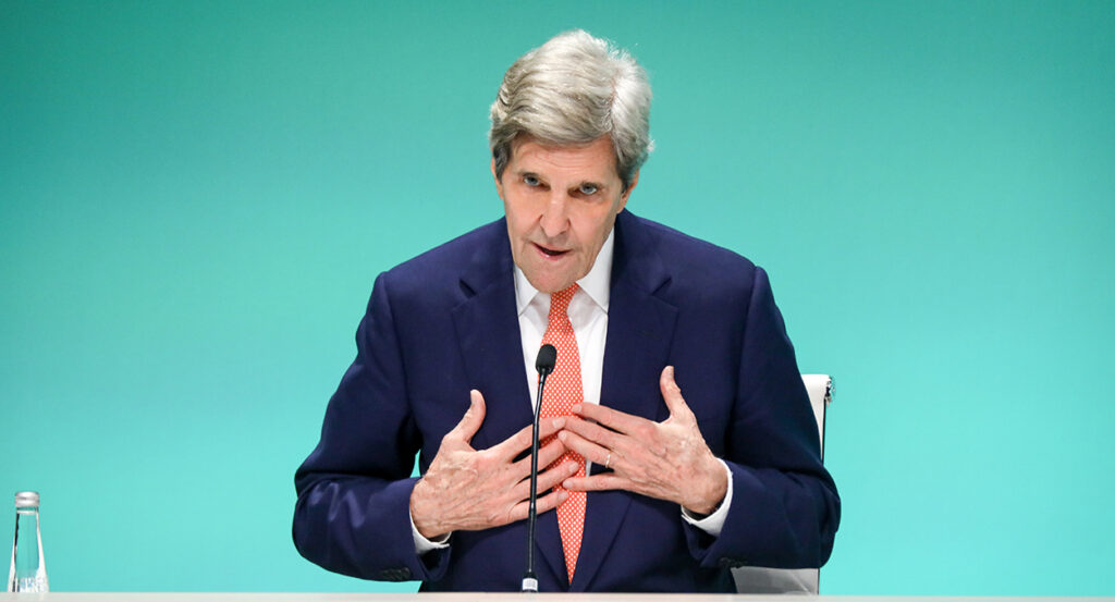 John Kerry in a blue suit gestures toward himself
