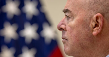 Side profile view shows DHS Secretary Alejandro Mayorkas looking ahead with an American flag behind him.
