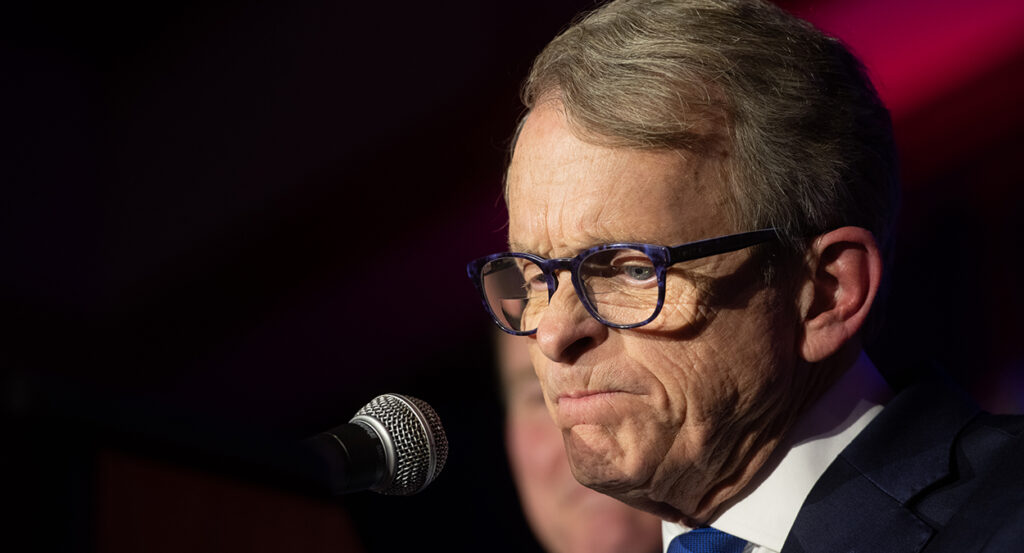 Ohio Governor Mike DeWine grimaces while wearing a suit in front of a microphone