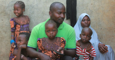 A Nigerian man sits with his wife and children outside their home