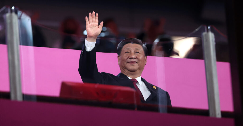 Chinese President Xi Jinping In a suit At a podium behind protective glass waves to an unseen crowd