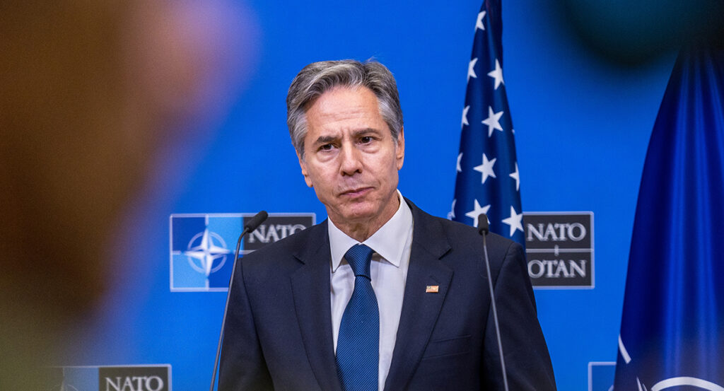 Antony Blinken in a blue suit purses his lips in front of an American flag and a NATO symbol.