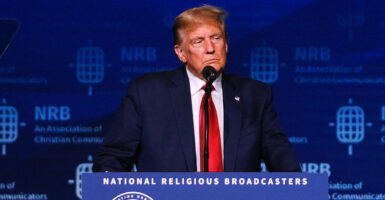 Donald Trump in a blue suit with a red tie stands behind a podium reading "National Religious Broadcasters"